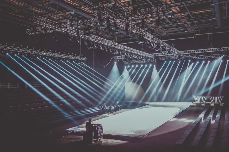 Empty stage in a large venue, lit by beams from an overhead rig of moving lights, with a technician at the lighting desk