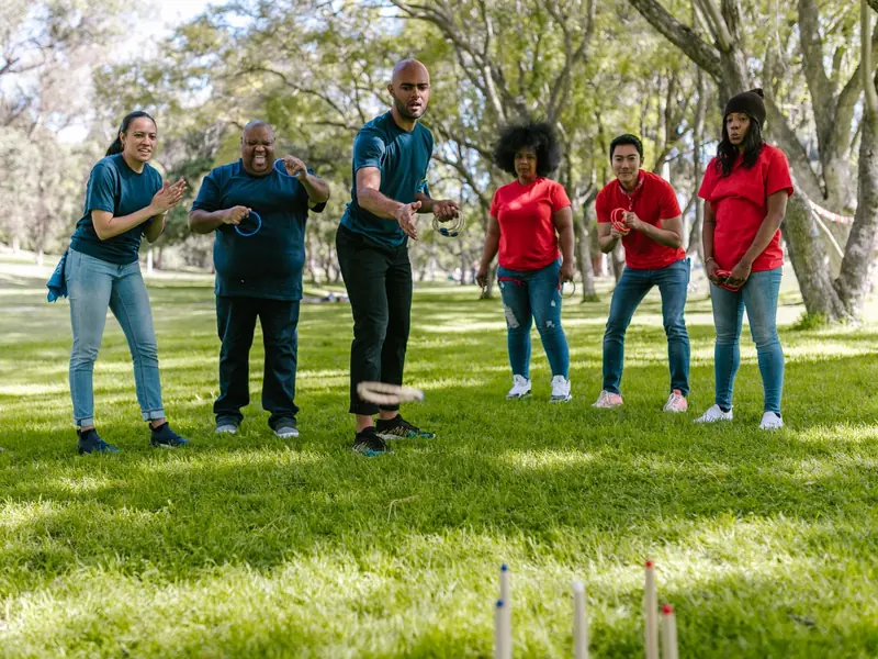 Six people in blue and red t-shirts playing a ring-toss game on a lawn under trees