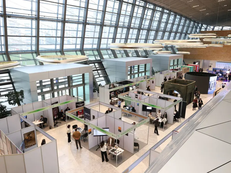 Exhibition hall seen from a gallery, with rows of white stand booths and visitors under a glass roof